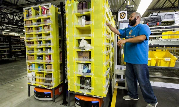 „Amazon“ sandėlyje du robotai (apačioje, oranžiniai) atvežė iki darbuotojo stelažus su užsakytais produktais. Bendrovės nuotr.